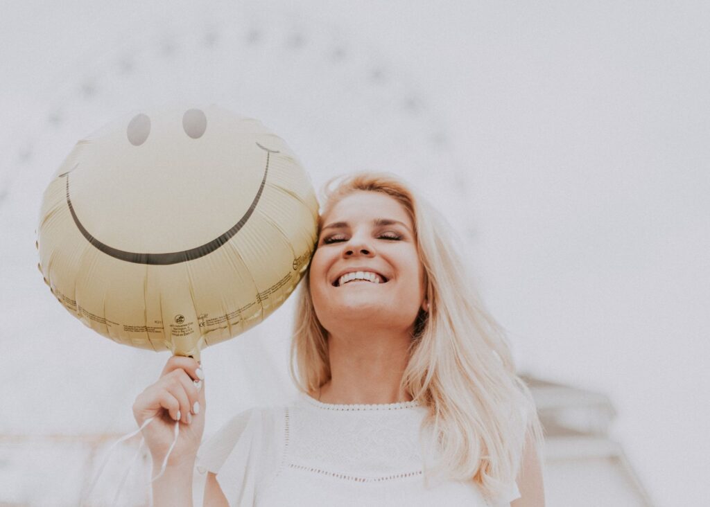 Blonde woman smiling next to a happy face balloon, looking like an expert in how to have more positive thoughts