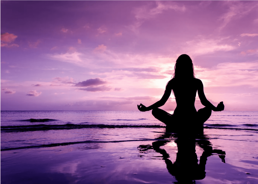 Silhouette of a woman engaged in meditation in front of a purple sunset.