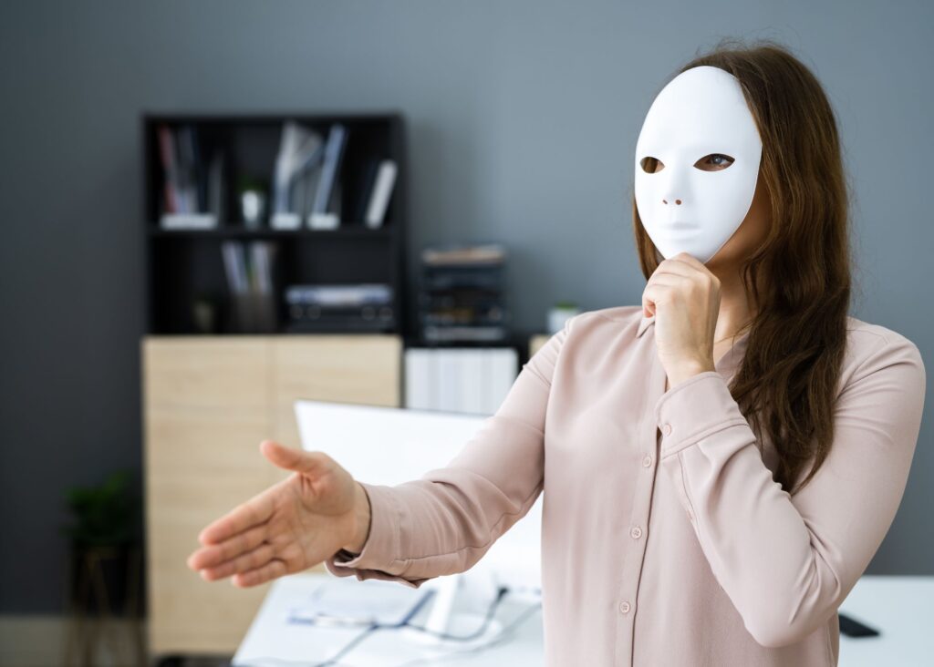 A brunette holding out her hand for a handshake and covering her face with a mask indicative of her imposter syndrome