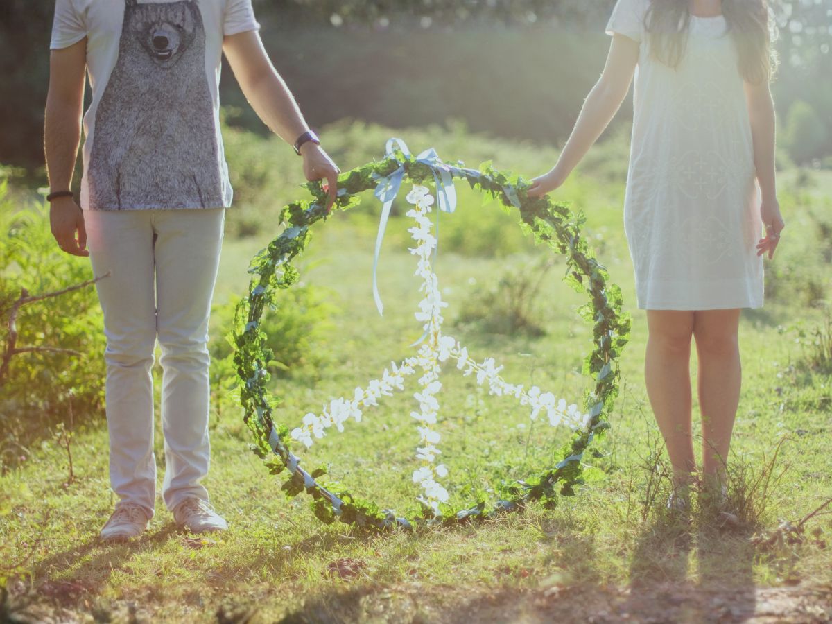 Claude responded: Two people standing in a sunlit field, holding between them a large circular wreath made of green leaves and vines, with white flowers arranged inside it in th…Two people standing in a sunlit field, holding between them a large circular wreath made of green leaves and vines, with white flowers arranged inside it in the shape of a peace sign.
