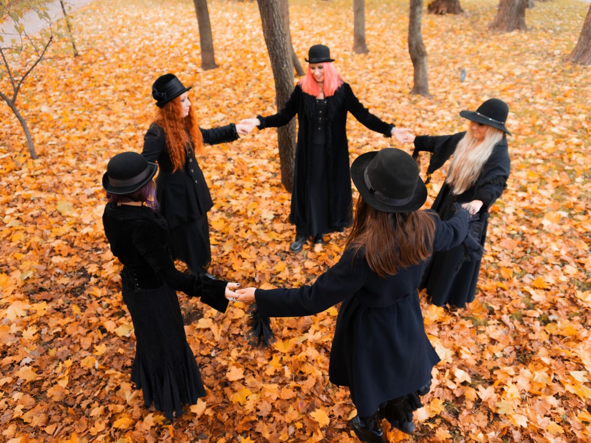 Five women in black witch attire and bowler hats stand in a circle holding hands on a forest floor covered in golden autumn leaves.